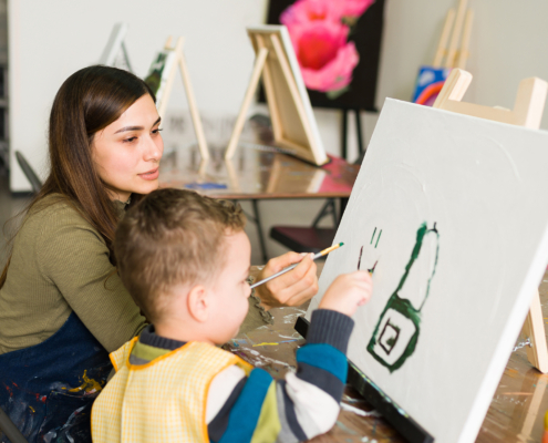 Young teacher and a kid student with aprons are painting on a canvas